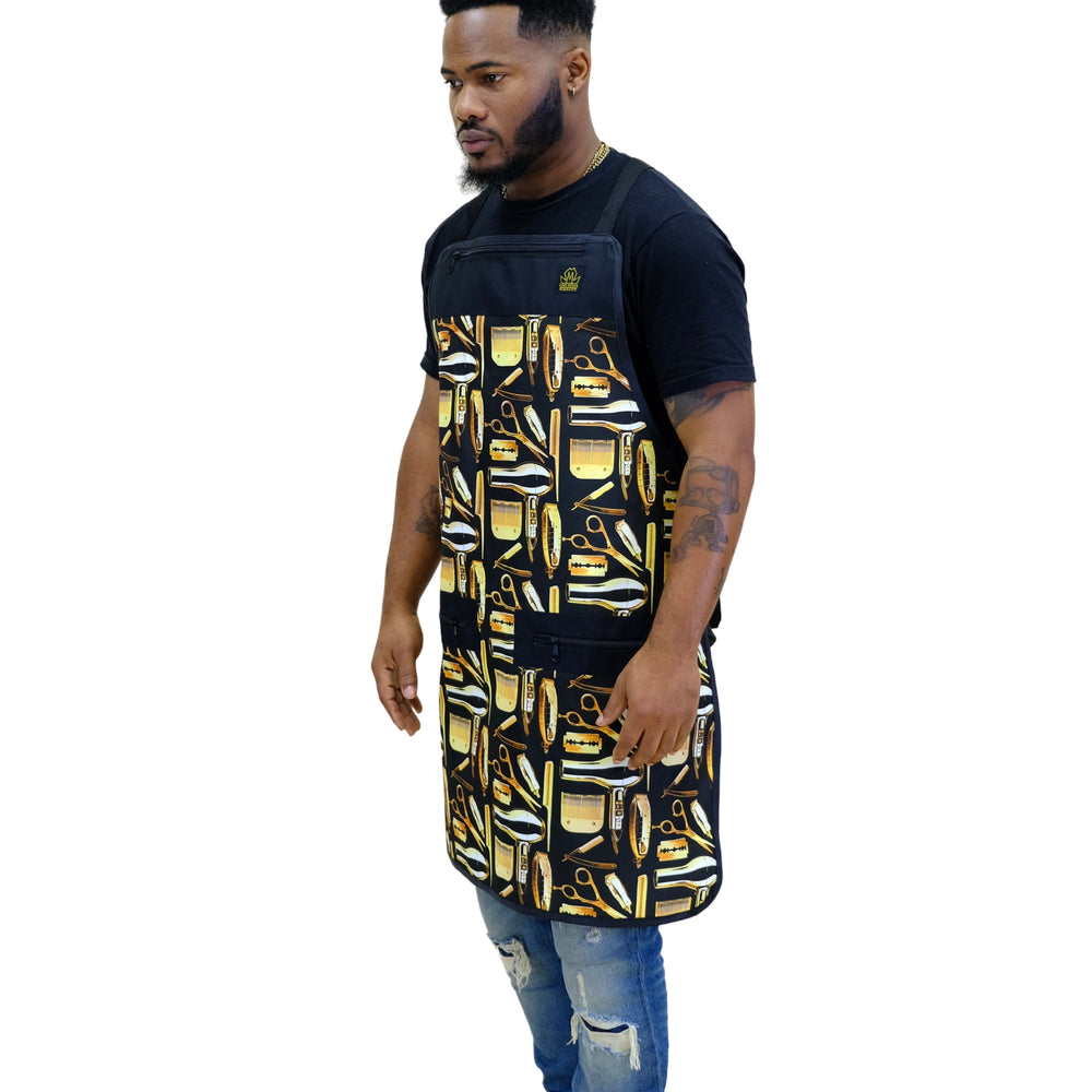 A man wearing a black barber apron with a yellow and white barber tools pattern, styled over a black t-shirt and blue jeans, shown against a plain white background.