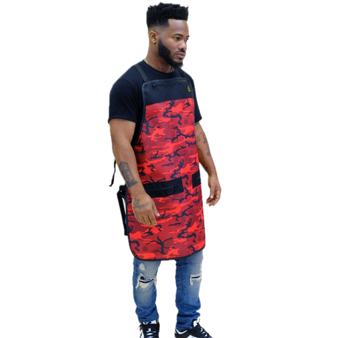 A man wearing a red and black camouflage barber apron over a black t-shirt and blue jeans, standing against a plain white background.