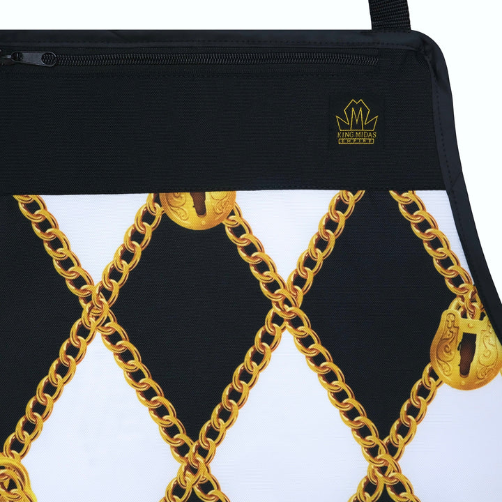 A close-up of a black zippered pouch on a barber apron featuring a gold chain and padlock pattern on a white background, with a small gold crown logo on the black header.