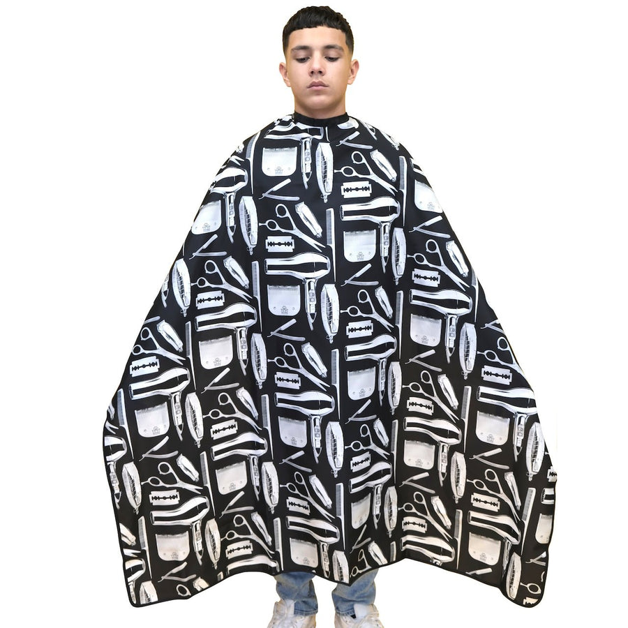 A  model wearing a black and white barber cape with a pattern of scissors, razors, and combs, standing against a solid white background.