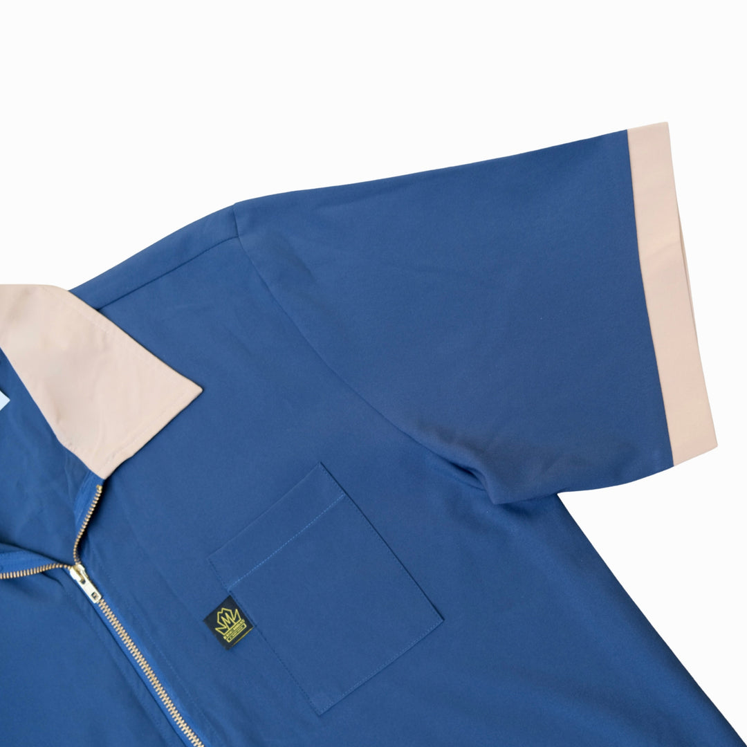 A close-up of a blue short-sleeve zip-up barber smock work shirt with a beige collar and sleeve cuffs, featuring a chest pocket and a small embroidered logo patch.