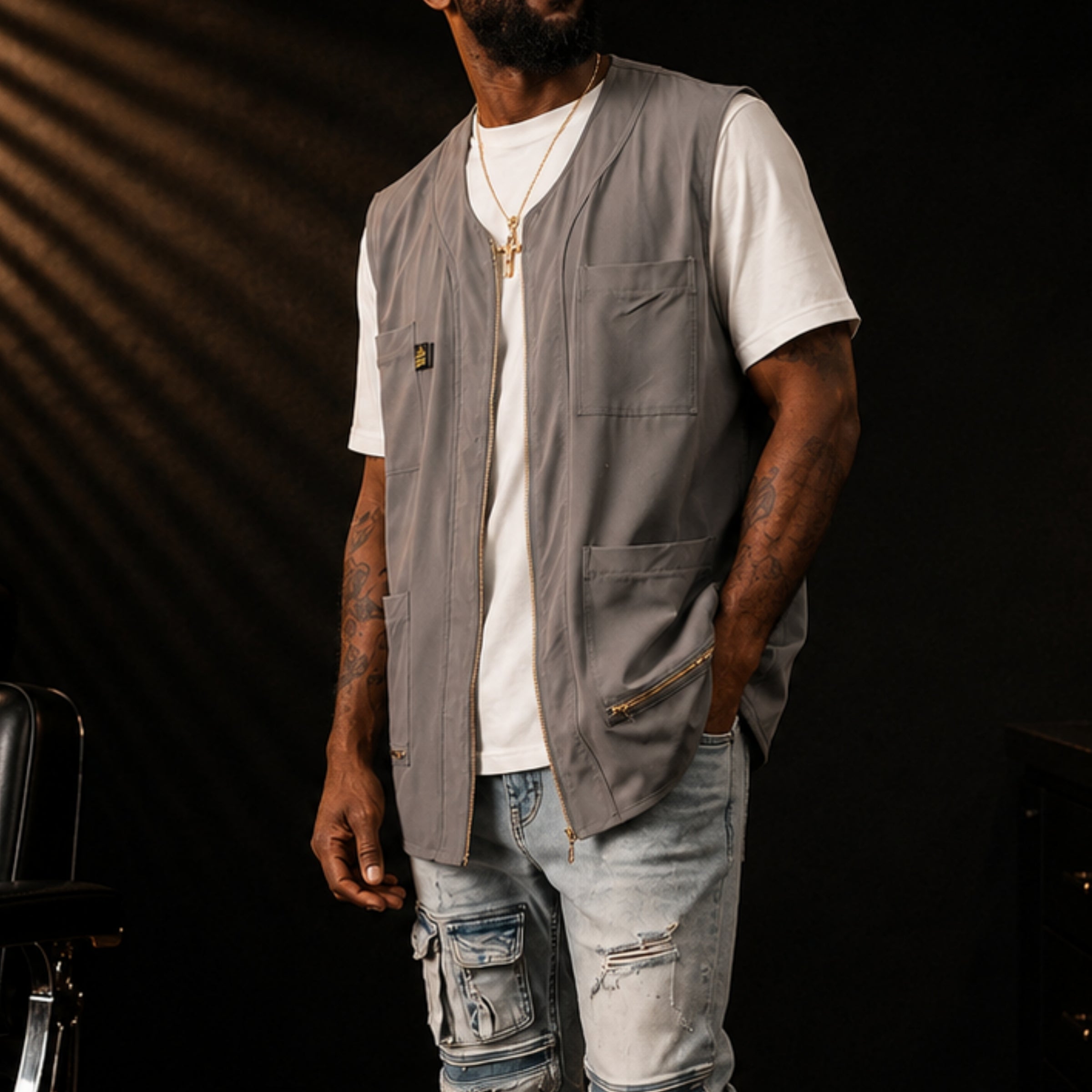 A man wearing a  gray zippered barber vest over a white t-shirt and light-wash distressed jeans, standing against a dark background with dramatic diagonal lighting.
