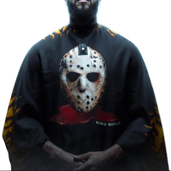A man wearing a black barber cape featuring a large graphic of a Jason hockey mask with blood splatter and the text 'KING MIDAS' on the front, with yellow flame-like graphics on the sleeves, set against a plain white background.