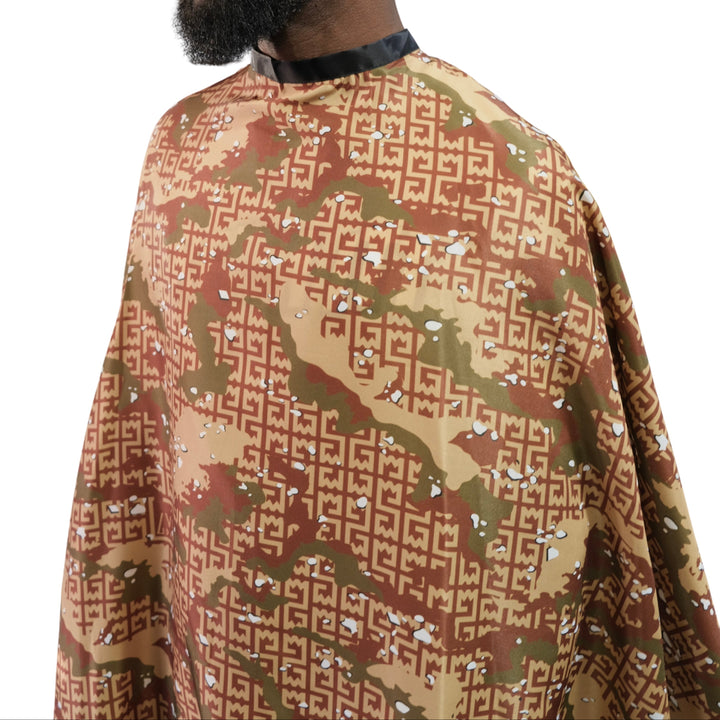 A close-up side view of a person wearing a  barber cape. The cape features a camouflage-style design in brown, tan, and olive green, overlaid with a repeating interlocking monogram pattern.