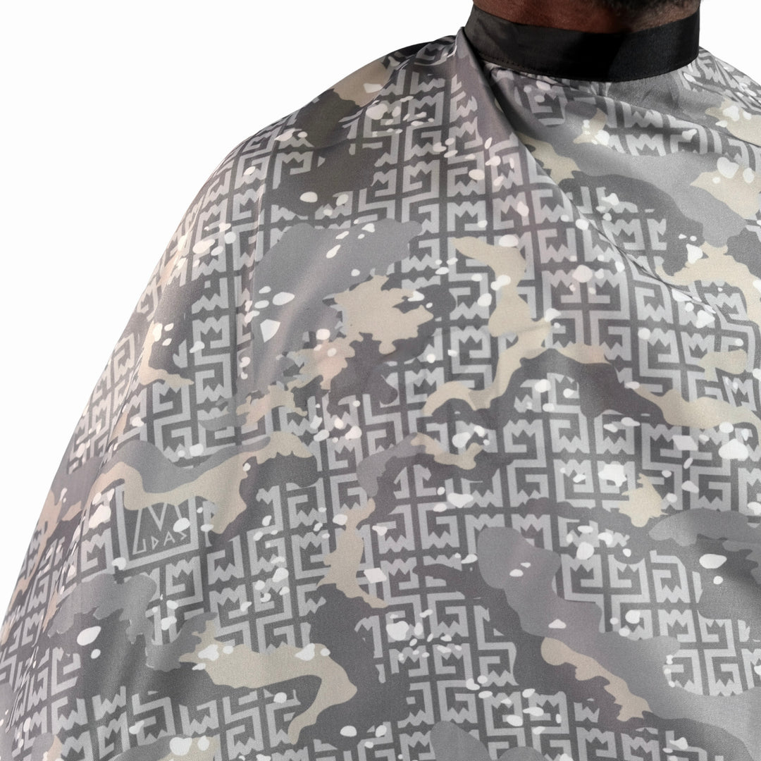 Close-up of a barber cape with a gray, beige, and white geometric camouflage pattern, worn by a person whose neck and collar are visible at the top.