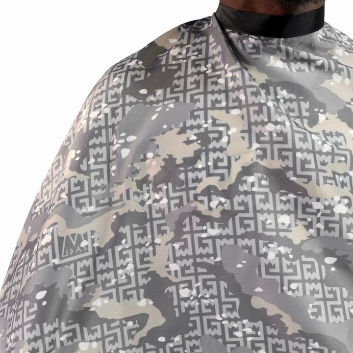 Close-up of a barber cape with a gray, beige, and white geometric camouflage pattern, worn by a person whose neck and collar are visible at the top.