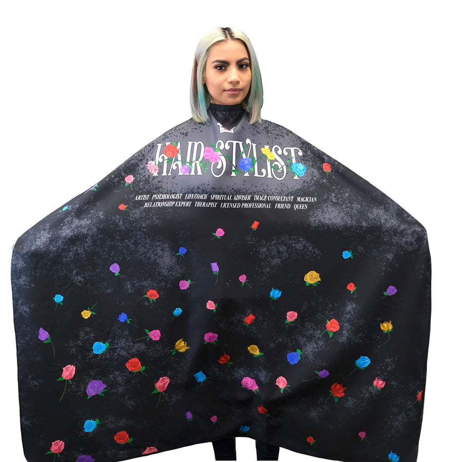 A person wearing a large black salon cape with a floral print and the words 'HAIR STYLIST' across the chest, isolated on a white background.